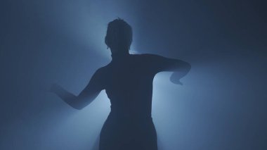 Işık konseptinde siluet. Stüdyoda siyah dumanlı arka planda parlak arka plana karşı kadın modeli. Kadın silueti, hareket eden el karanlıkta bir ışık demeti ile oynuyor..