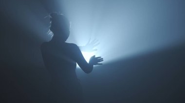 Işık konseptinde siluet. Stüdyoda siyah dumanlı arka planda parlak arka plana karşı kadın modeli. Kadın silueti, hareket eden el karanlıkta bir ışık demeti ile oynuyor..