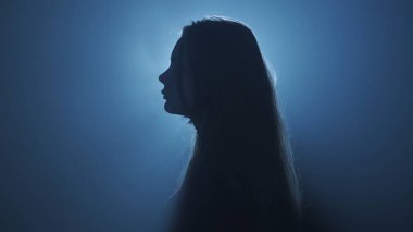Işık konseptinde siluet. Stüdyoda siyah dumanlı arka planda parlak ışığa karşı kadın modeli. Karanlıkta ışık huzmeleriyle çevrili bir kadın silueti. Yan görünüm