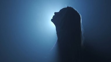 Işık konseptinde siluet. Stüdyoda siyah dumanlı arka planda parlak ışığa karşı kadın modeli. Karanlıkta ışık huzmeleriyle çevrili bir kadın silueti. Yan görünüm