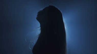 Işık konseptinde siluet. Stüdyoda siyah dumanlı arka planda parlak ışığa karşı kadın modeli. Karanlıkta ışık huzmeleriyle çevrili bir kadın silueti. Yan görünüm