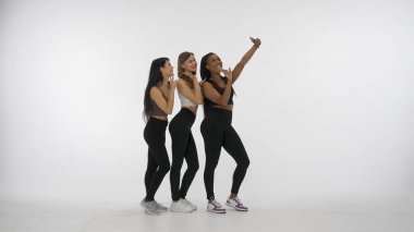 Beyaz stüdyo arka planında izole edilmiş genç çoklu etnik modellerin portresi. Akıllı telefondan selfie çeken üç pozitif çok ırklı kız grubu. Çok ırklı güzellik kavramı