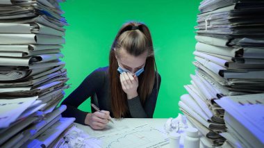 Sıkı çalışma konsepti. Yorgun, genç bir iş kadını ofis çalışanı gece geç saatlere kadar evrak işleriyle meşgul. Yüz maskeli soğuk algınlığı olan hasta bir kadın masa başında kağıtlarla çalışıyor. Yeşil