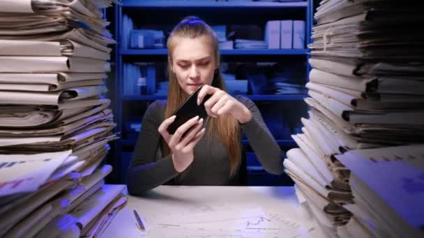 努力工作的概念 疲惫不堪的年轻商业女性办公室职员深夜在办公桌前工作 文件堆积如山 女性在智能手机上玩游戏拖延时间 — 图库视频影像