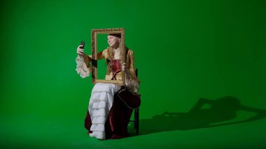 Tarihsel kişi modern yaşam tarzı reklamı. Krom anahtar yeşil ekran arka planda antik kıyafetli bir kadın. Rönesans tarzı elbise giymiş bir kadın resim çerçevesi tutuyor ve akıllı telefondan selfie çekiyor..