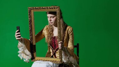 Tarihsel kişi modern yaşam tarzı reklamı. Krom anahtar yeşil ekran arka planda antik kıyafetli bir kadın. Rönesans tarzı elbise giymiş bir kadın resim çerçevesi tutuyor ve akıllı telefondan selfie çekiyor..