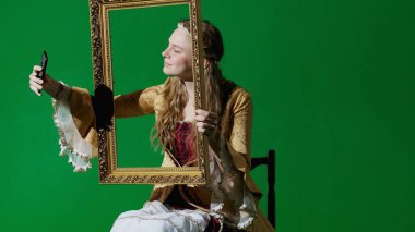 Tarihsel kişi modern yaşam tarzı reklamı. Krom anahtar yeşil ekran arka planda antik kıyafetli bir kadın. Rönesans tarzı elbise giymiş bir kadın resim çerçevesi tutuyor ve akıllı telefondan selfie çekiyor..