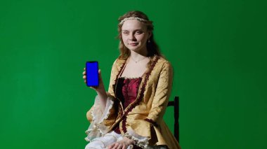 Tarihsel kişi modern yaşam tarzı reklamı. Krom anahtar yeşil ekran arka planda antik kıyafetli bir kadın. Rönesans elbisesi giymiş, elinde akıllı telefon ve çalışma alanı modeli olan bir kadın baş parmağını gösteriyor..