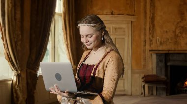 Tarihsel kişi modern yaşam tarzı reklamı. Tarihi iç mekanın arka planında, kadim kıyafetli bir kadın. Rönesans tarzı elbise giymiş bir kadın dizüstü bilgisayarda mesaj yazıyor..