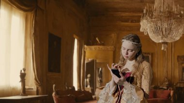 Tarihsel kişi modern yaşam tarzı reklamı. Tarihi iç mekanın arka planında, kadim kıyafetli bir kadın. Rönesans tarzı elbiseli bir kadın akıllı telefon tutuyor ve mesaj yazıyor.
