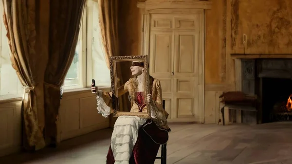 Historical man modern lifestyle advertising. Woman in ancient outfit on background of historic interior. Woman in historical dress holding a framed picture and taking selfies on smartphone