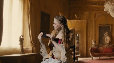 Tarihsel kişi modern yaşam tarzı reklamı. Tarihi iç mekanın arka planında, kadim kıyafetli bir kadın. Rönesans tarzı elbise giymiş bir kadın elinde çikolatayla oturmuş, şeker yiyor..