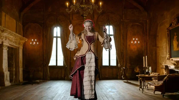 Tarihsel kişi modern yaşam tarzı reklamı. Tarihi iç mekanın arka planında, kadim kıyafetli bir kadın. Rönesans tarzı giyinen, kulaklıklı bir kadın akıllı telefondan müzik dinliyor ve dans ediyor..