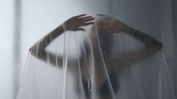Body beauty and fashion advertisement. Female in underwear sitting on the bed in the studio. Young woman seminude in lingerie sits posing with thin sheet of oilcloth wrap, looks at camera.