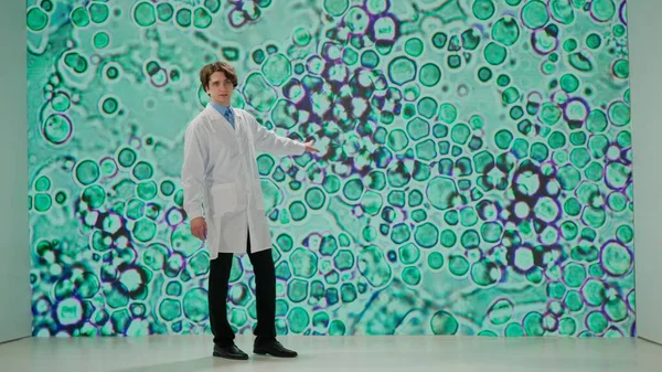実験室のコートの科学者は 拡大された細胞構造を表示する大きなデジタルスクリーンの前に立って 専門的または学術的な設定で複雑な生物学的概念を説明します — ストック写真