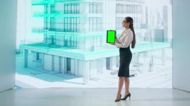 Bir kadın, çok katlı bir binanın holografik projeksiyonuyla etkileşime girer ve yeşil ekran tableti gösterir. Dijital ekran, mekanı bir projenin estetiğiyle aydınlatıyor.