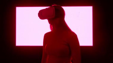 Dijital görsel teknoloji kavramı. Sanal gerçeklik gözlüklü bir kadın silueti dijital ekran duvarının önünde duruyor ve etrafta video oyunu oynayan neon semboller var..