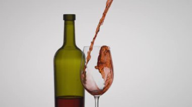 Kırmızı şarabı şeffaf bir şarap bardağına doldurmanın dinamik anı. Beyaz arka plan parlak şarap ve yeşilimsi boş şişeyle çelişiyor. Kapat..