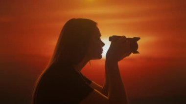 Gün batımının parlak gölgesinde kamerayla fotoğraf çeken genç bir kadının silueti..
