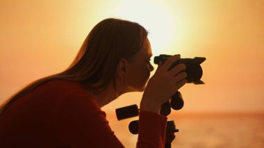 Bu resimde, altın saat boyunca fotoğraf çeken bir kadın fotoğrafçının silueti yer alıyor. Arka plandaki sıcak günbatımı yaratıcılığın huzurlu anını vurgular..
