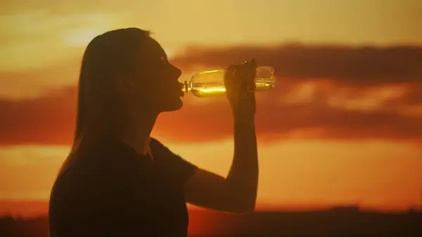 The silhouette of a person enjoying a refreshing drink from a bottle is captured against the warm glow of a setting sun. The tranquil scene evokes a sense of peace and the simple pleasure of hydration