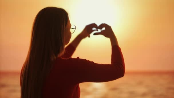 在这个视频中 一个女人在日落背景下的轮廓使她的心形围绕着太阳 她的双手象征着爱和与自然的联系 — 图库视频影像