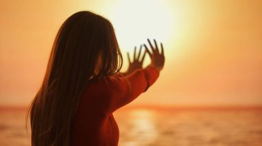Görüntü huzurlu bir anı yakalar. Bir kadın batan güneşe dokunmak için uzanır ve canlı günbatımının arka planına karşı neşeli bir siluet yaratır.