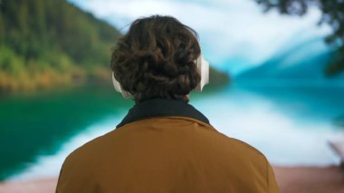 Genç adam büyük kablosuz kulaklıkla müzik dinliyor. Yeşil dağları ve gölü olan bulanık bir manzaraya karşı sıcak giysiler içinde rahat bir tatil atmosferi taşıyan bir adam. Arka plan.