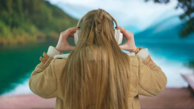 Geniş kablosuz kulaklıkla müzik dinleyen genç bir kadın. Yeşil dağları ve gölü olan bulanık bir manzaraya karşı sıcak giysiler içinde rahat bir tatil atmosferi taşıyan bir kadın. Arka plan.