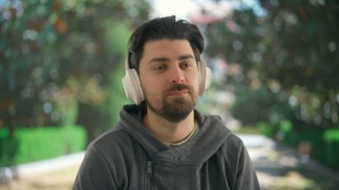Geniş kablosuz kulaklıkla müzik dinleyen genç bir adam. Bulanık bir park manzarasının önündeki yeşil ağaçlı erkek dinlenme atmosferini taşır..
