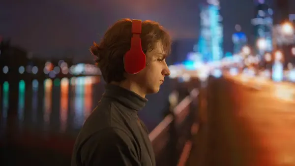 Young man in profile, listening to music with vibrant red headphones against a backdrop of a blurry cityscape at night, with the shimmering lights of the urban skyline reflected on the waters surface