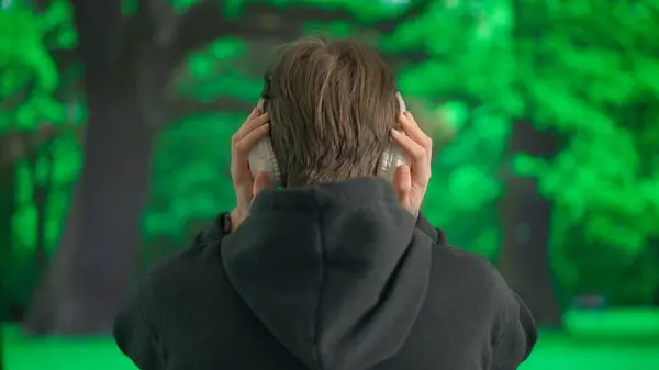 一个年轻人在大的无线耳机里听音乐 男人在一片绿树成荫的公园前 传达着一种悠闲的休息氛围 背景色 — 图库照片