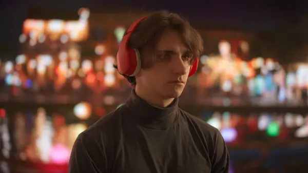 Young man in profile, listening to music with vibrant red headphones against a backdrop of a blurry cityscape at night, with the shimmering lights of the urban skyline reflected on the waters surface