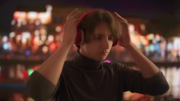 プロフィールの若い男は 夜のぼやけた街並みを背景に鮮やかな赤いヘッドフォンで音楽を聴き 都市のスカイラインの輝く光が水面に映し出された — ストック動画