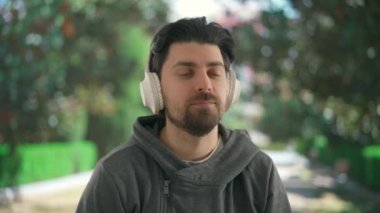 Geniş kablosuz kulaklıkla müzik dinleyen genç bir adam. Bulanık bir park manzarasının önündeki yeşil ağaçlı erkek dinlenme atmosferini taşır..