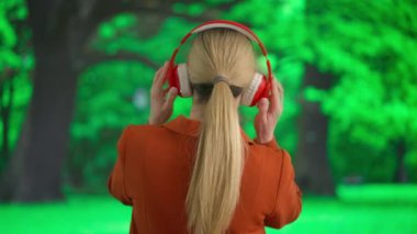 Geniş kablosuz kulaklıkla müzik dinleyen genç bir kadın. Bulanık bir park manzarasının önünde yeşil ağaçlarla rahatlamış bir dinlenme atmosferi taşıyan bir kadın. Arka plan.