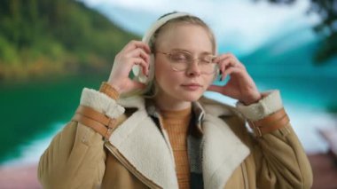 Geniş kablosuz kulaklıkla müzik dinleyen genç bir kadın. Yeşil dağları ve gölü olan bulanık bir manzaraya karşı sıcak giysiler içindeki bir kadın rahat bir tatil atmosferini taşıyor..