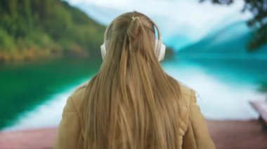 Geniş kablosuz kulaklıkla müzik dinleyen genç bir kadın. Yeşil dağları ve gölü olan bulanık bir manzaraya karşı sıcak giysiler içinde rahat bir tatil atmosferi taşıyan bir kadın. Arka plan.