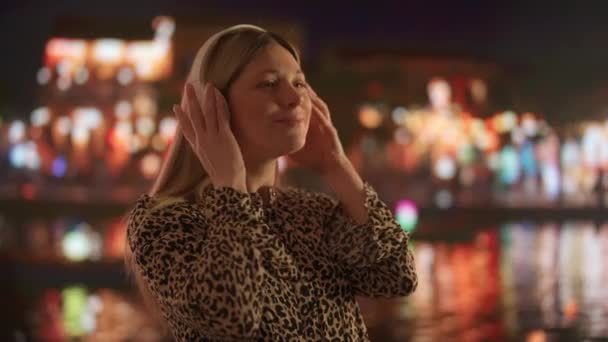 大きなホワイトヘッドホンで音楽を聴く若い女性 夜の明るい街並みを背景に 都市のスカイラインの輝く光が水面に映し出される — ストック動画