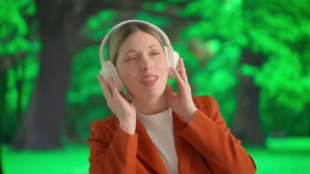 一个年轻的女人用大的无线耳机听音乐 在一片绿树成荫的公园前 女性的身影闪烁着 营造出一种悠闲的休息氛围 — 图库视频影像