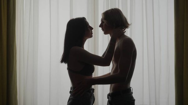 Love intimate relationships. Young loving couple silhouettes in apartment. Man and woman half naked sensual lovers standing against window together, passion and intimacy kissing.
