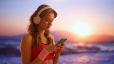 Gadget 'lar ve modern günlük hayat reklamları konsepti. Her gün akıllı telefon kullanan bir kadın. Gün batımında plajda kulaklıklarla dans eden, akıllı telefondan müzik dinleyen genç bir kadın..