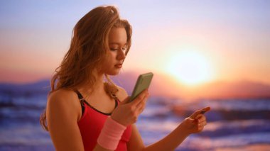 Gadget 'lar ve modern günlük hayat reklamları konsepti. Her gün akıllı telefon kullanan bir kadın. Gün batımında plajda spor kıyafetleri içinde genç bir kadın elinde akıllı bir telefonla uygulamayla kredi kartı numarasını yazıyor..