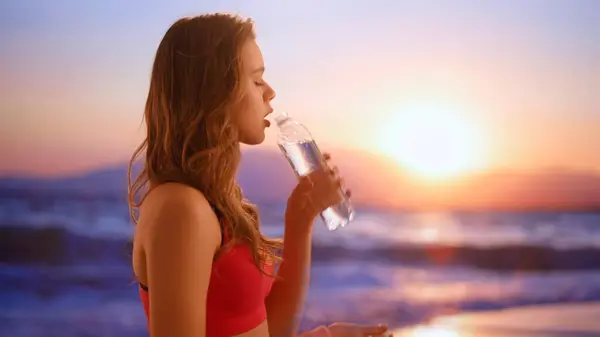 Health and body wellness in everyday life advertisement concept. Female model on the beach at sunset. Young athletic woman in sportswear on the beach drinking water from a bottle.
