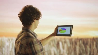 Tarım ve modern teknoloji kavramı. Gün batımında büyük buğday tarlasına karşı çiftçi. Tarımcı adam tarım alanında duruyor, elinde tablet tutuyor ürün büyümesini kontrol ediyor, mali sonuçları ekranda