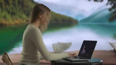 Serbest ve serbest meslek hayatı konsepti. Uzaktan çalışan ve seyahat eden bir kadın. Dağ Gölü yakınlarında çalışan, dizüstü bilgisayarda yazan, elinde kağıt belgeler tutan serbest çalışan genç bir kadın..