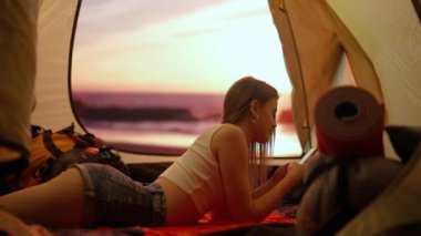 Kamp ve macera konsepti. Kamp alanında gezen, yürüyüş yapan, doğayı keşfeden biri. Kumsalda gün batımında çadırın içinde akıllı telefondan mesajlaşan genç bir kadın. Tatil ve açık havada tatil.