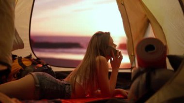 Kamp ve macera konsepti. Kamp alanında gezen, yürüyüş yapan, doğayı keşfeden biri. Genç bir kadın, gün batımında sahilde çadırın içinde akıllı telefondan konuşuyor. Tatil ve tatilde dışarıda.