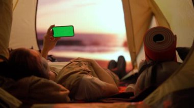 Kamp ve macera konsepti. Kamp alanında gezen, yürüyüş yapan, doğayı keşfeden biri. Akıllı telefon krom anahtar yeşil ekranlı genç adam kumsaldaki çadırın içinde, dışarıda tatil yapıyor..
