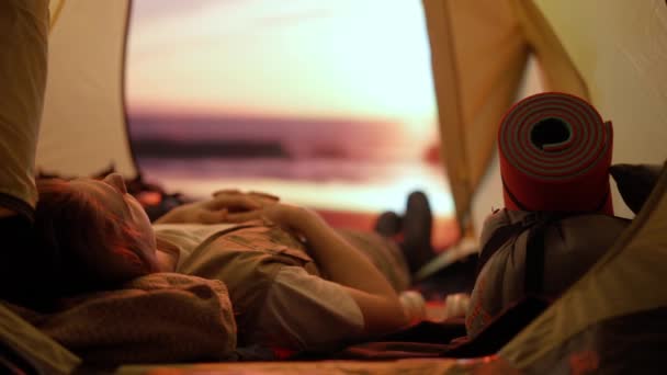 キャンプと冒険のコンセプト キャンプ場の旅行やハイキング 自然を探検する人 ビーチの日没のテントの内側のスマートフォンで音楽を聞くヘッドフォンに横たわる若い男 — ストック動画